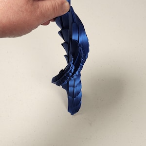 Puede incluir: Una escultura abstracta azul oscuro impresa en 3D con un diseño en capas en forma de ala. El objeto está sostenido por una mano y parece estar hecho de un material brillante. La escultura tiene una forma curva y una superficie texturizada.