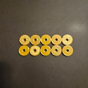 May include: Ten small, gold-colored, circular objects with a hole in the center are arranged in two rows of five on a dark surface. The objects have a textured surface and a metallic sheen.