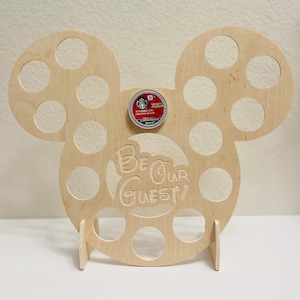 May include: A wooden coffee pod holder shaped like Mickey Mouse's head with 16 holes for coffee pods. The holder has a stand and the text "Be Our Guest!" is engraved on the front.