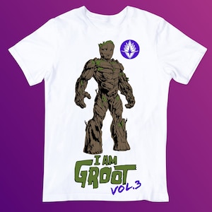 Può includere: T-shirt bianca con un'illustrazione di Groot dei Guardiani della Galassia. Il design include il testo "I AM GROOT VOL. 3" in verde e blu. Un emblema circolare è presente anche sulla maglietta.