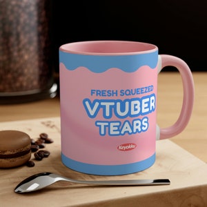 Könnte beinhalten: Eine rosa Keramiktasse mit blauem Rand und weißem Henkel. Die Tasse hat einen rosa Hintergrund mit blauem Text, der "Fresh Squeezed VTuber Tears" lautet, und ein rotes Logo mit der Aufschrift "Keyokku".