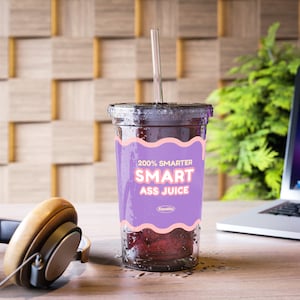 Könnte beinhalten: Ein durchsichtiger Plastikbecher mit einem lila und pinkfarbenen Etikett, auf dem "200% Smarter Smart Ass Juice" steht, mit einem Strohhalm, der aus der Oberseite herausragt. Der Becher ist mit einer dunkelroten Flüssigkeit gefüllt und hat Kondensation an der Außenseite. Der Becher steht auf einer Holzoberfläche.