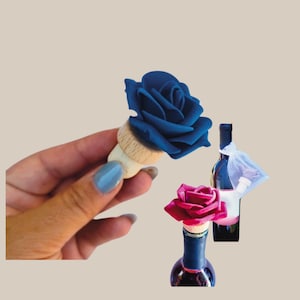 Può includere: Tappi decorativi per bottiglie di vino con rose. Un tappo presenta una rosa blu scuro, mentre un altro mostra una rosa magenta. Viene mostrata anche una bottiglia di vino con un tappo bianco e un sacchetto di rete bianco.
