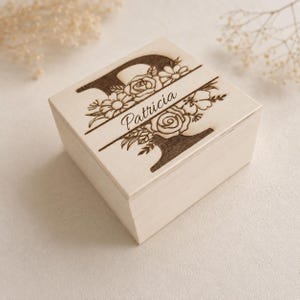 May include: A square light-colored wooden box featuring a laser-engraved design on the lid. The design includes a large letter P, floral illustrations, and the name Patricia written in a cursive script across the center of the letter.