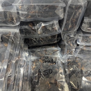 May include: Multiple clear plastic resealable bags filled with dark brown dried snails. Several bags feature handwritten black ink text that reads Dry Snails and $50. The contents appear to be small, curled, and dehydrated organic specimens.