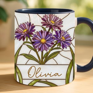 May include: White ceramic mug with a stained-glass floral design featuring purple and pink flowers with yellow centers. The mug has a navy blue handle and rim, and the name "Olivia" is written in gold script.