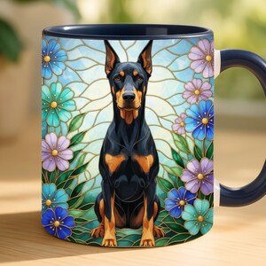 May include: A ceramic mug with a dark blue handle and interior. The mug features a Doberman Pinscher illustration surrounded by colorful flowers. The dog is black and tan, set against a stained-glass style background with blue, green, and purple flowers.