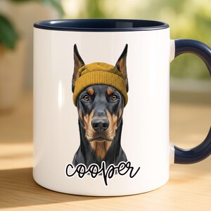 May include: White ceramic mug with a navy blue handle and rim. The mug features a portrait of a Doberman Pinscher wearing a yellow beanie. The dog's name, "Cooper", is written in cursive below the portrait.