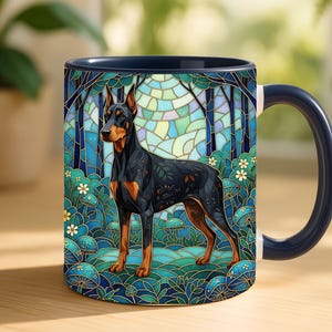 May include: A ceramic mug featuring a Doberman Pinscher illustration in a stained-glass style. The dog is black and tan, set against a blue and green forest scene. The mug has a dark blue handle and rim, and is set on a wooden surface.