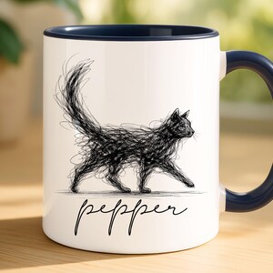 May include: White ceramic mug with a navy blue handle and rim. The mug features a black scribble drawing of a cat walking, with the name "pepper" written below in a script font. The mug is on a wooden surface.