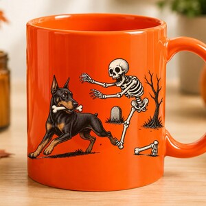 May include: A bright orange ceramic mug featuring a graphic illustration of a black and tan Doberman dog holding a white bone in its mouth while being chased by a white human skeleton near a small tombstone and a bare tree silhouette.