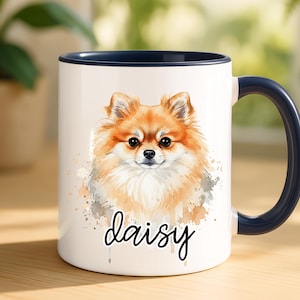 May include: White ceramic mug with a navy blue handle and rim. The mug features a watercolor illustration of a Pomeranian dog with orange and white fur. The dog's name, "daisy", is written in cursive below the image.