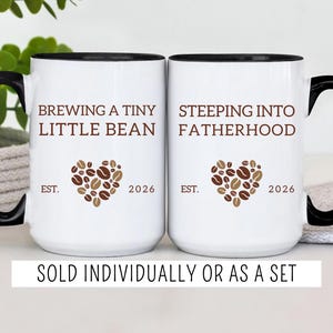 May include: Two white ceramic mugs with black handles and rims. The left mug features the brown text "BREWING A TINY LITTLE BEAN" above a heart made of coffee beans and "EST. 2026". The right mug features "STEEPING INTO FATHERHOOD" with a heart.