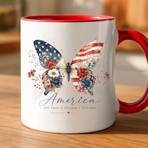 May include: White ceramic mug with a red interior and handle. The mug features a butterfly design with the American flag on the wings, floral accents, and the word "America" with the text "250 Years of Freedom 1776-2026".