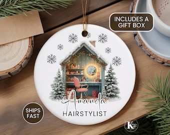 May include: A round white ceramic ornament featuring a graphic of a hair salon interior with a pink chair, shelves of products, and a round mirror. The scene is framed by two snow-covered pine trees and snowflakes. Text reads Amanda HAIRSTYLIST.