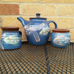 Puede incluir: Una tetera azul y dos tazas con un diseño de pescado. La tetera tiene un asa y una tapa marrones. Las tazas tienen asas marrones. Los peces están pintados en rosa, azul y amarillo.