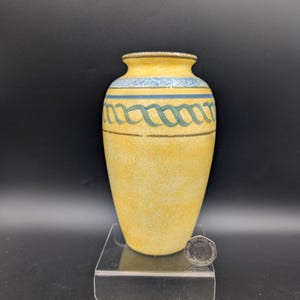 May include: A yellow ceramic vase with a textured surface. The vase features a blue band near the top and a green, intertwined design. The vase is approximately 6 inches tall.