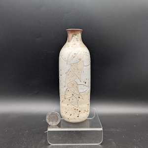 Op de afbeelding: Een cilindrische keramische fles met een smalle hals en een gespikkeld crèmekleurig glazuur. Het lichaam heeft een ingesneden bladpatroon en een donkerbruine rand. Een Britse munt van twintig pence ligt naast de voet en meet 2,14 cm.