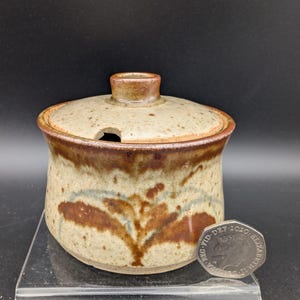 Peut inclure: Un sucrier en céramique avec couvercle, présentant un glaçage beige et marron moucheté. Le bol a un motif floral marron et une petite ouverture pour une cuillère. Le couvercle a une petite poignée.