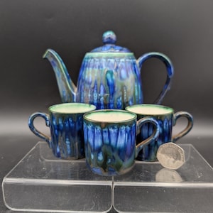 May include: A ceramic teapot with a lid and three matching mugs, all featuring a glossy blue and green drip glaze finish. The interior of the mugs is a light cream colour. A small silver-coloured coin is placed next to the mugs for scale.