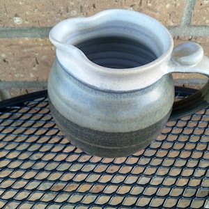 May include: A grey and white ceramic jug with a handle and a rounded spout. The jug has a black band around the bottom and a white rim.