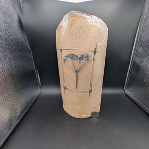 Könnte beinhalten: Eine beige Keramikvase mit floralem Design. Die Vase hat eine raue, strukturierte Oberfläche und eine leicht unregelmäßige Form. Das florale Design ist in einem rechteckigen Rahmen mit vertikalen Linien eingeschlossen. Die Vase ist etwa 25 cm hoch.