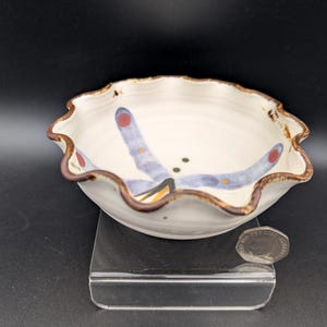 May include: A white ceramic bowl with a scalloped, brown-rimmed edge featuring a painted blue dragonfly design with red and orange accents. A silver-colored coin is placed next to the bowl for scale, providing a clear visual reference for its size.