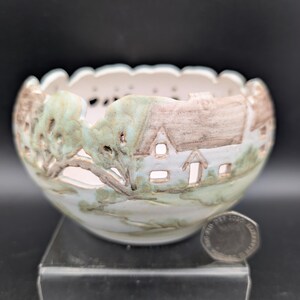 May include: A white ceramic bowl featuring a cut-out design of a landscape with houses and trees in muted brown and green tones. The bowl has a scalloped rim and is positioned next to a British twenty pence coin for scale, showing its size.