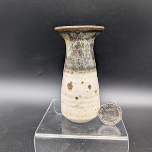 May include: A small ceramic vase with a flared rim, featuring a speckled cream-colored base that transitions into a dark, mottled grey-brown glaze at the top. A British fifty-pence coin is placed next to the vase for scale reference.