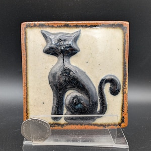 Puede incluir: Azulejo cerámico cuadrado con un gato negro. El gato está sobre un fondo crema con un borde marrón. La forma del gato está en relieve, creando un efecto 3D. El azulejo se exhibe en un soporte transparente.