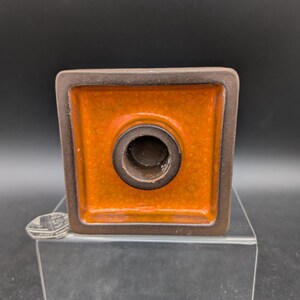 May include: A square ceramic candle holder with a brown rim and a bright orange interior. The candle holder has a circular opening in the center and sits on a clear acrylic stand. The background is black.