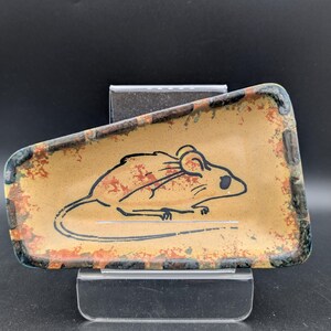 May include: A rectangular, decorative dish with a tan background and a black outline of a mouse. The dish has a distressed, rustic look with brown and red accents around the edges. The dish is displayed on a clear stand.