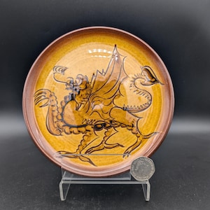 Può includere: Un piatto in ceramica con un disegno di drago. Il piatto ha un bordo marrone e un centro giallo con un'illustrazione dettagliata di un drago in marrone e nero. Il drago ha le ali ed è in una posa dinamica.