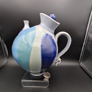 Può includere: Una teiera in ceramica fatta a mano con un corpo bulboso e un manico curvo. La teiera presenta uno schema di colori a strisce blu, bianche e turchese. Ha un piccolo beccuccio e un coperchio con un pomello blu. La teiera è esposta su un supporto trasparente.