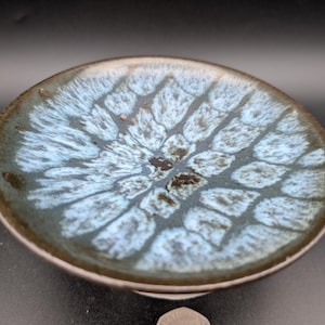 May include: A small, round ceramic dish with a unique glaze. The interior features a light blue and white pattern, while the rim is a darker, earthy brown. The dish has a slightly raised edge and a smooth, glossy finish. The overall aesthetic is organic and artistic.