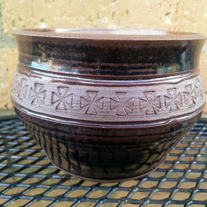 May include: A brown ceramic pot with a decorative band of cross-shaped designs. The pot has a dark brown glaze and a light brown band.