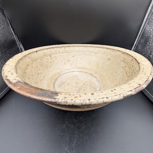 May include: A speckled, beige ceramic bowl with a wide rim. The rim has a dark brown band around the edge. The bowl's interior has a slightly raised centre. This handmade bowl is suitable for serving or display.
