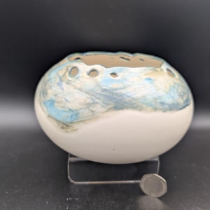 Puede incluir: Un cuenco de cerámica blanco y redondo con una abertura superior ancha que presenta una serie de pequeños recortes circulares a lo largo del borde. La parte superior del cuenco está decorada con un patrón marmoleado texturizado en tonos azul claro y gris.