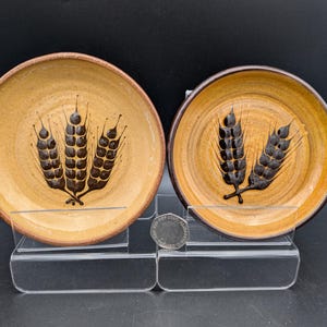 May include: Two small, round, ceramic dishes with a wheat stalk design. The dishes have a tan base with a brown rim and a dark brown wheat stalk design. The dishes are displayed on a clear acrylic stand against a black background.