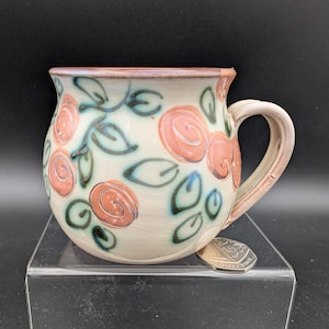 May include: A ceramic mug with a cream-colored base, adorned with hand-painted pink roses and green leaves. The mug has a rounded shape, a sturdy handle, and a rim with a reddish-brown hue. A coin rests on the surface next to the mug.