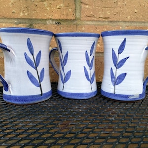 Può includere: Tre tazze in ceramica fatte a mano con corpi bianchi, bordi e manici blu. Ogni tazza presenta un disegno a foglie blu. I manici sono anch'essi blu, alcuni con strisce. Le tazze sono esposte su una superficie metallica nera.