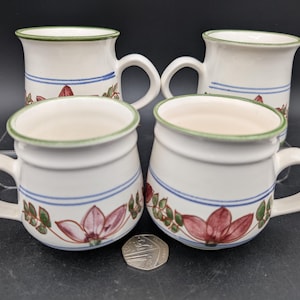May include: Four white ceramic mugs with a floral design. Each mug has a green rim, a blue stripe, and a pink and red flower with green leaves. The mugs have a curved handle. The mugs are on a dark surface.