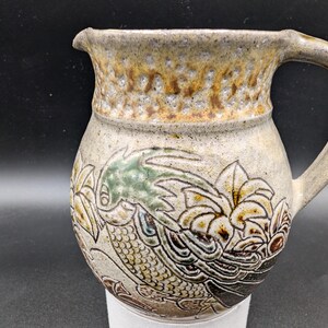 Könnte beinhalten: Eine Keramikkanne mit brauner, weißer und grüner Glasur. Die Kanne hat ein Blumenmuster mit einem Vogel an der Seite.