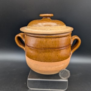 Può includere: Un vaso in ceramica a due manici con coperchio, caratterizzato da una smaltatura marrone lucida sulla parte superiore e una finitura opaca in terracotta sulla base inferiore. Una piccola moneta argentata è posta alla base come riferimento.