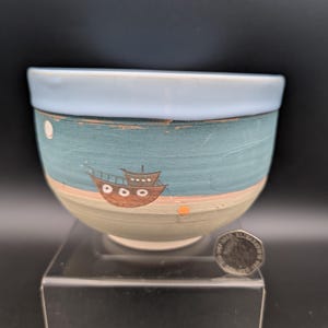 Könnte beinhalten: Handbemalte Keramikschale mit hellblauem Rand und Meereslandschaftsdesign. Die Schale hat einen türkisblauen Hintergrund mit einer braunen Bootsillustration, einem beigen Strand und einem hellblauen Himmel. Die Schale hat einen Durchmesser von ca. 10 cm.