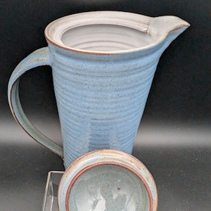 May include: A light blue ceramic jug with a ribbed, textured exterior and a smooth white interior. The jug features a curved handle on the side and a matching circular lid with a brown rim, shown resting in front of the main vessel.