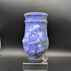 Może przedstawiać: Ceramiczny wazon o teksturowanej powierzchni z żywą fioletową glazurą nakrapianą ciemnoszarymi i czarnymi akcentami. Wazon ma rozszerzany brzeg i zaokrąglony korpus, stoi na przezroczystej akrylowej podstawie obok małej srebrnej monety dla skali.