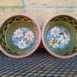 May include: Two brown ceramic bowls with a green glaze and a white, pink, and yellow floral design. The bowls are sitting on a black metal surface.