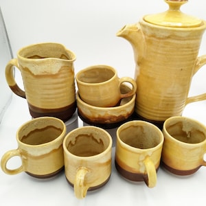 Pode incluir: Um conjunto de seis canecas de cerâmica amarela e marrom e um jarro combinando. As canecas e o jarro têm um padrão listrado.