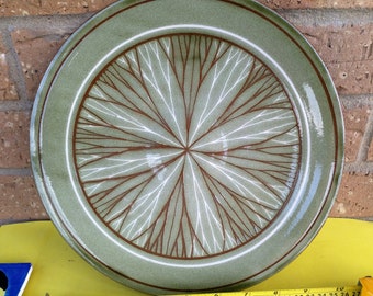May include: A green ceramic dinner plate with a white and brown geometric design. The design features a radial pattern of lines that resemble branches.